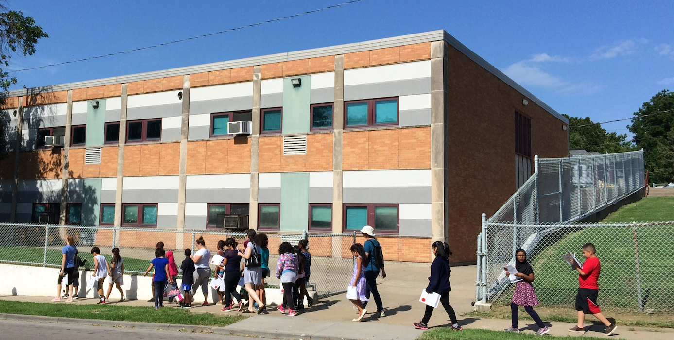 A group of students carrying workbooks walk down the sidewalk in front of their school. 